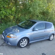 Błękitny Chevrolet Kalos SX, zaparkowany na kostce brukowej, na tle zielonych drzew.