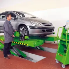 Mechanik sprawdza ustawienie kół samochodu na podnośniku. Obok stoi zielony wózek z komputerem, który pomoże określić, ile kosztuje ustawienie zbieżności.