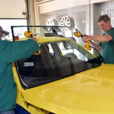 Dwóch mechaników wymienia przednią szybę w żółtym samochodzie. Dowiedz się, ile kosztuje wymiana szyby przedniej.