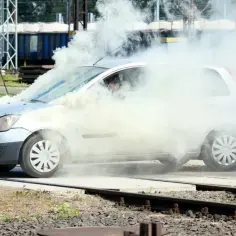 Srebrny samochód z kierowcą w środku, otoczony gęstym białym dymem, wygląda jak rozbiegany diesel na tle torów kolejowych.
