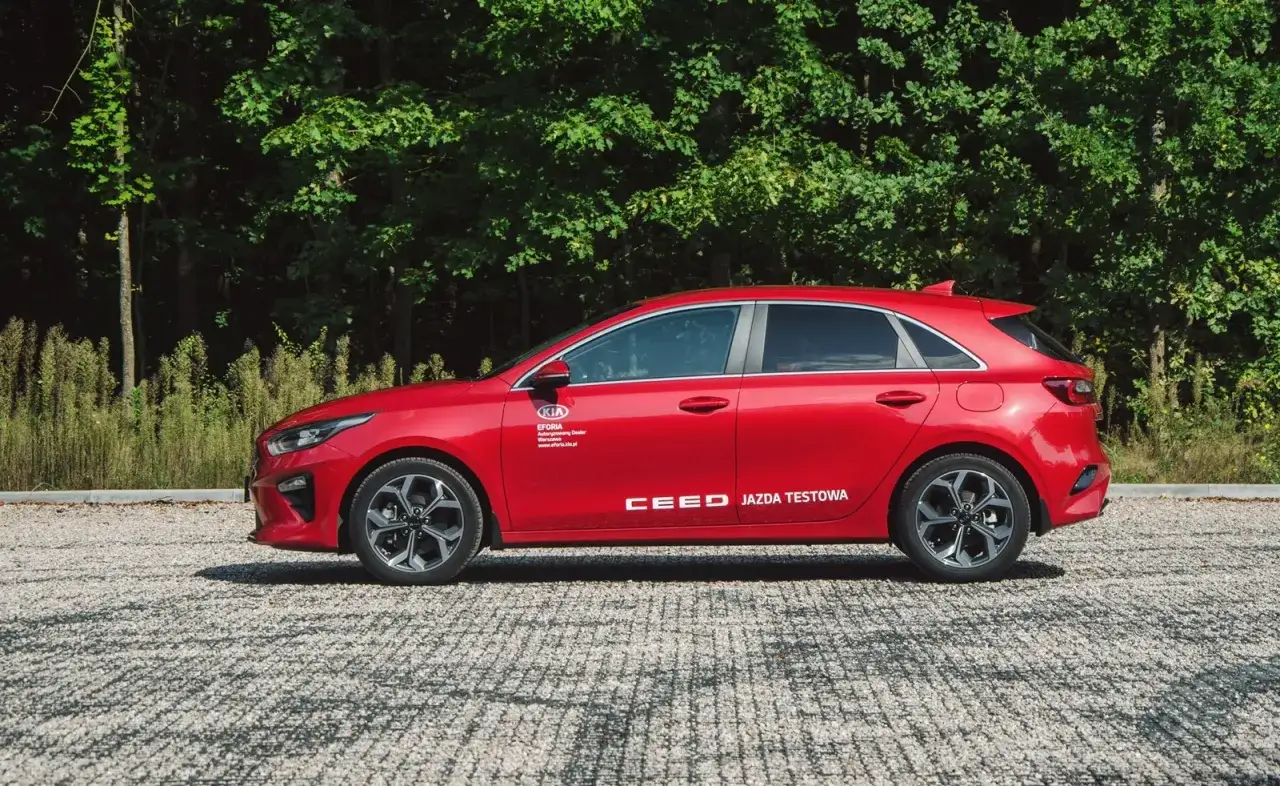 Czerwona Kia Ceed 1.4 benzyna, gotowa do jazdy testowej, zaparkowana na żwirowym podjeździe, z zielonym lasem w tle.
