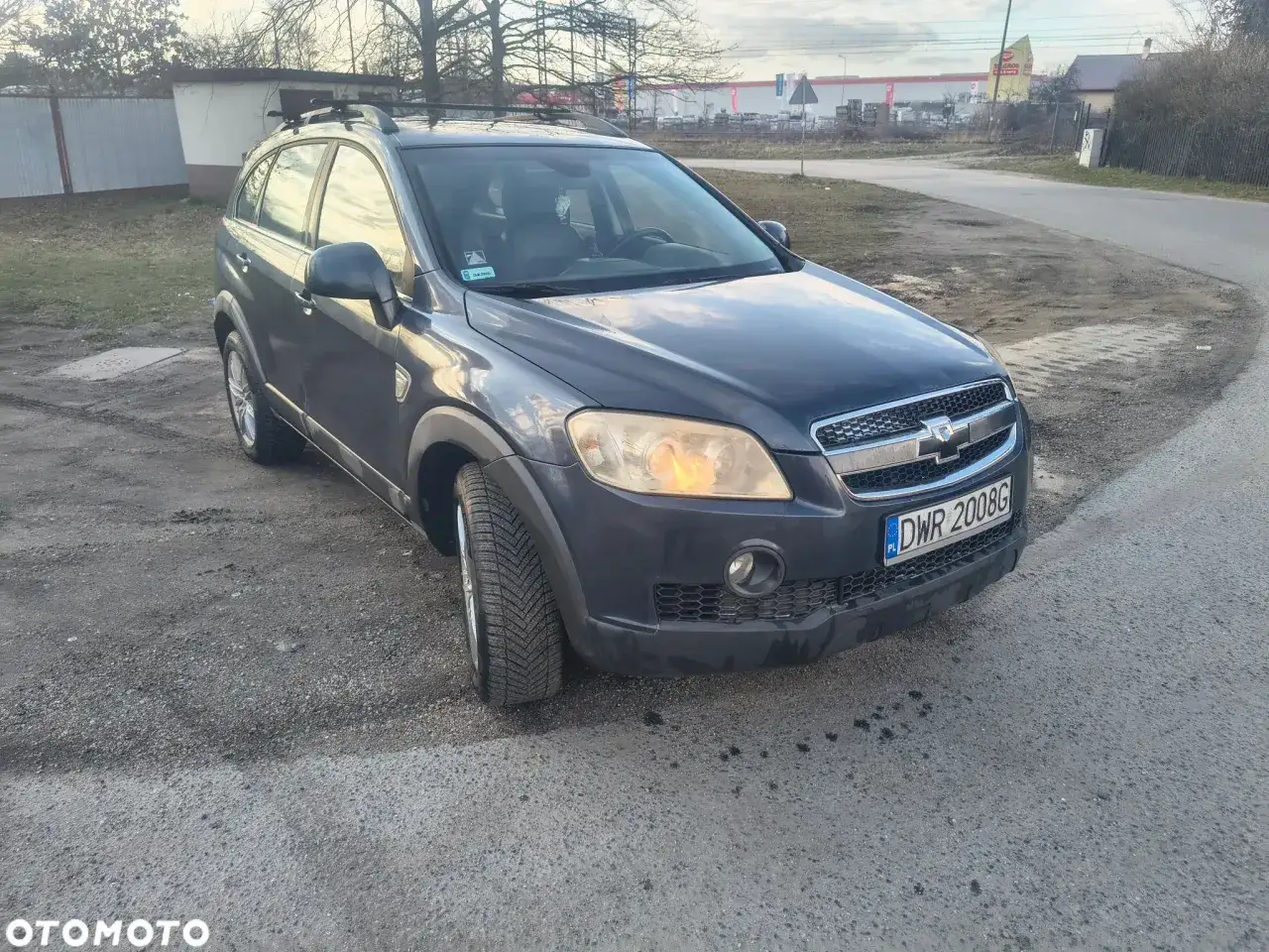 Ciemnoszary Chevrolet Captiva z relingami dachowymi stoi na szutrowej drodze.